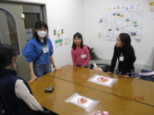 長机の端に立つ女性と、3名の女の子が本日の作業の確認をしている様子の写真