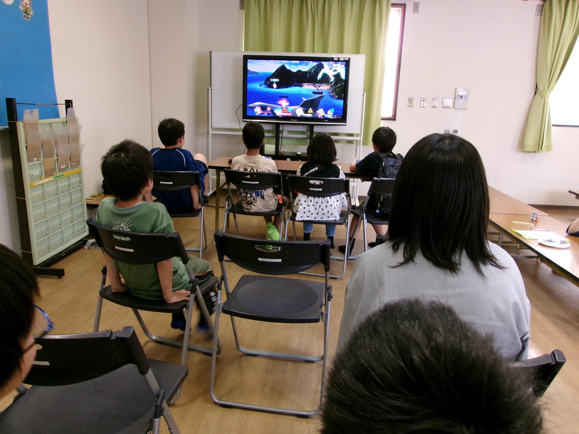 テレビ画面に映るゲームで遊んでいる子どもを観戦している子どもの写真