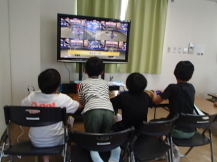 横並びに座った四人の子どもたちが、テレビモニターに映されるゲーム映像を見ながら真剣にゲームをしている写真