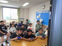 横並びに座って真剣にゲームをする数人の子どもたちと、そのゲームを観覧する子どもたちを、ななめ前から写した写真