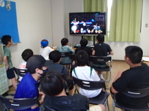 テレビモニターに映されるゲーム映像を見ながら真剣にゲームをする子どもたちと、その子どもたちの後ろに座って同じモニターを見る子どもたちの写真