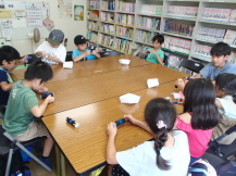 大きな机を囲むように座った子どもたちが、二つのフィルムケースを繋げて、そのケースにシールを貼っている写真