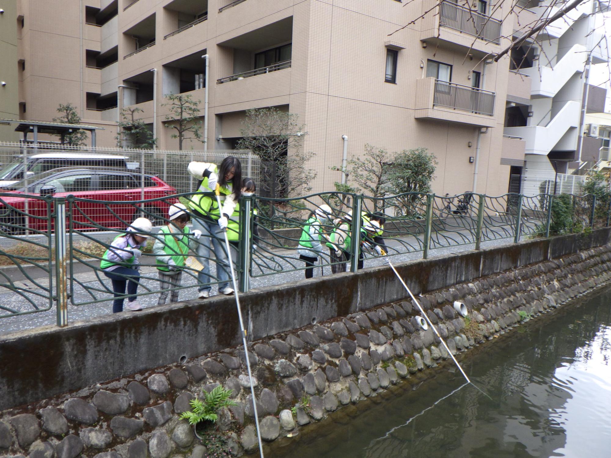 長い棒を使って川の中のゴミを高校生と一緒に拾っている子どもたちの写真