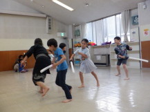 前の人を追いかけあって棒を振る4人の子供たちと先生の写真