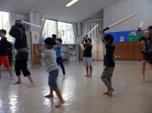 等間隔に2列に並び両手で持った棒を上から振りかざし素振りをしている子供たちと指導する先生の写真