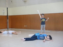 2人の子供が床にだいのじと体を横にして倒れているマネをしており勝った男の子が棒を片手にガッツポーズをしている写真