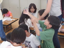 完成したオニヤンマを片手で持ち斜め下から眺める男の子と周りで黙々と作業する子供たちの写真
