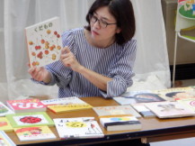 机に並べられた絵本の中の1冊を持つもっちゃん先生を撮影した写真