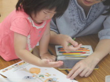 机に体を乗り出して紙に手形スタンプを押そうとしている女の子と横で紙を抑える先生の写真