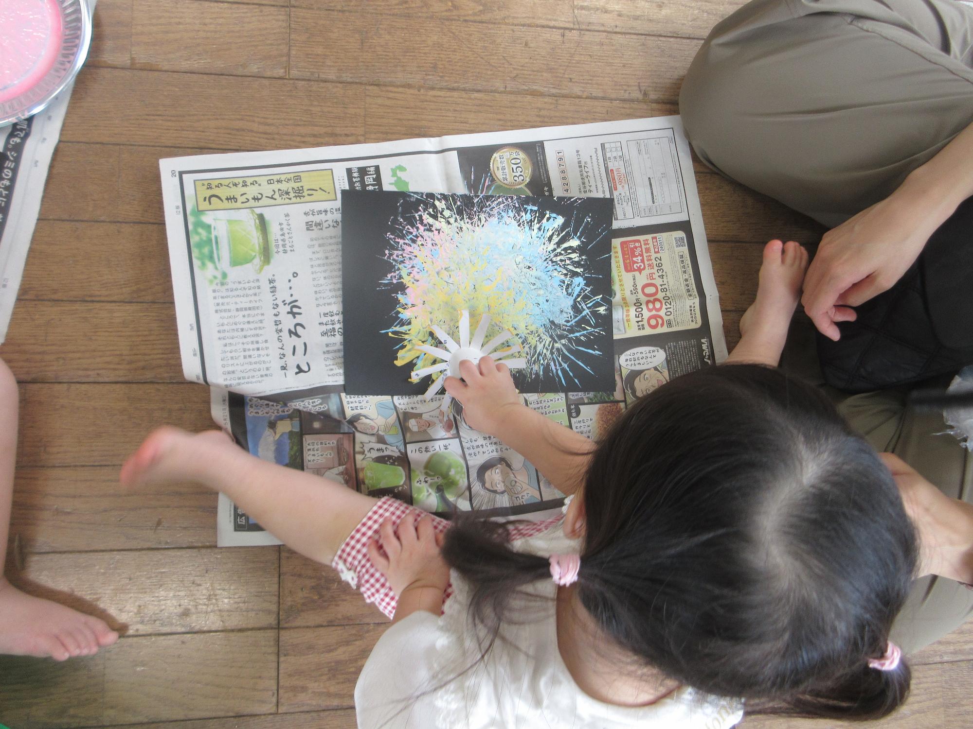 幼児がトイレットペーパーの芯の先を数本に切り開いたものに絵具をつけたものを使って、黒い色画用紙に花火の絵を描いているようす