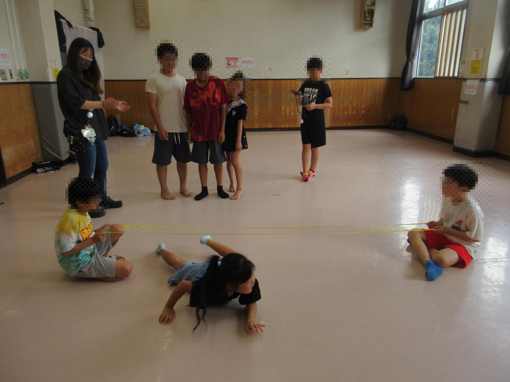 2名の児童が持って床から30センチほどの高さに張られた二本のリボンの下をくぐる児童と、それを見守るスタッフと児童たちのようす