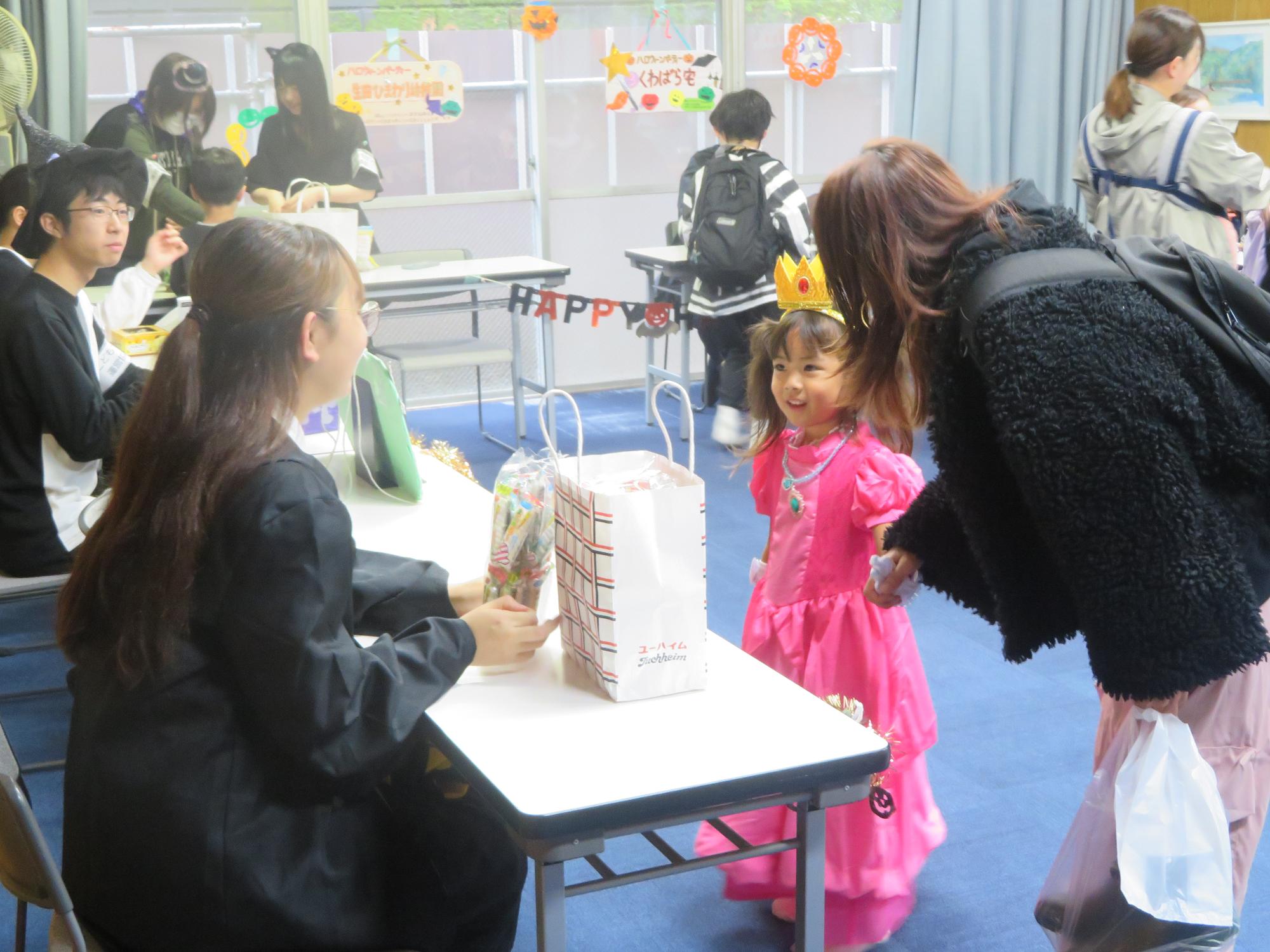 ピンクのドレスを着た幼児が映画キャラクターの仮装をしたボランティアからお菓子をもらっている写真