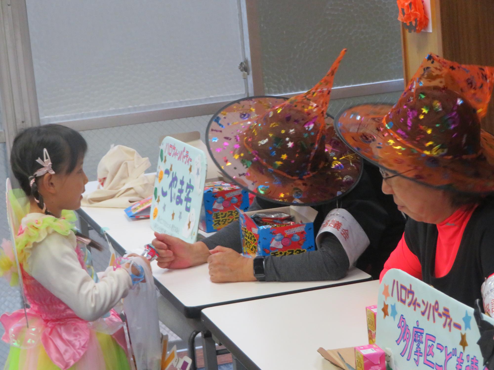 妖精の仮装をしたこどもがハロウィンの帽子を被ったボランティアからお菓子をもらっている写真