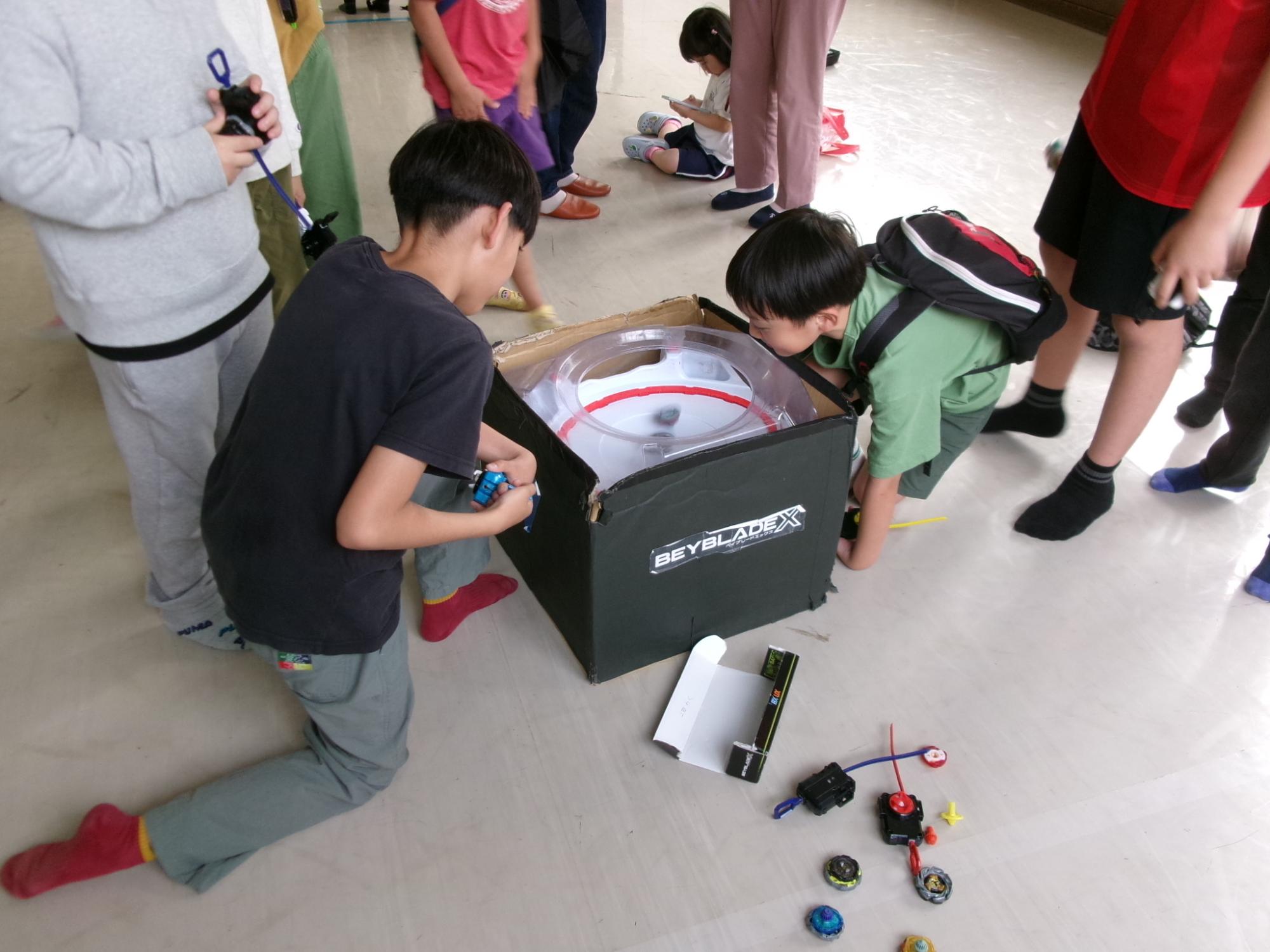 小学生児童がベイブレードの大会に熱中しまわりでその様子を子どもたちが取り囲んでいる写真
