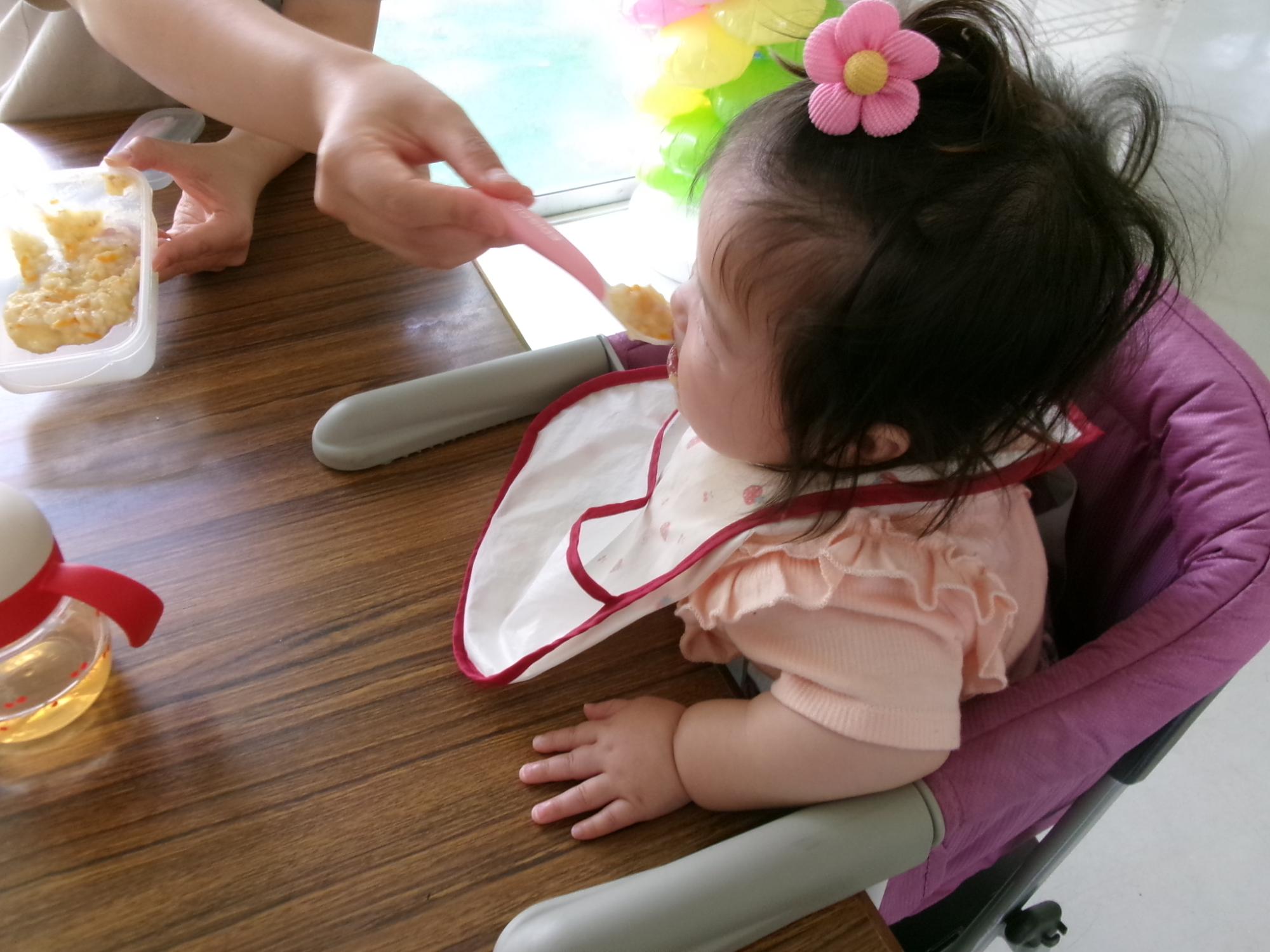 乳幼児がベビーチェアに座って母親から離乳食を食べさせてもらっている様子の写真