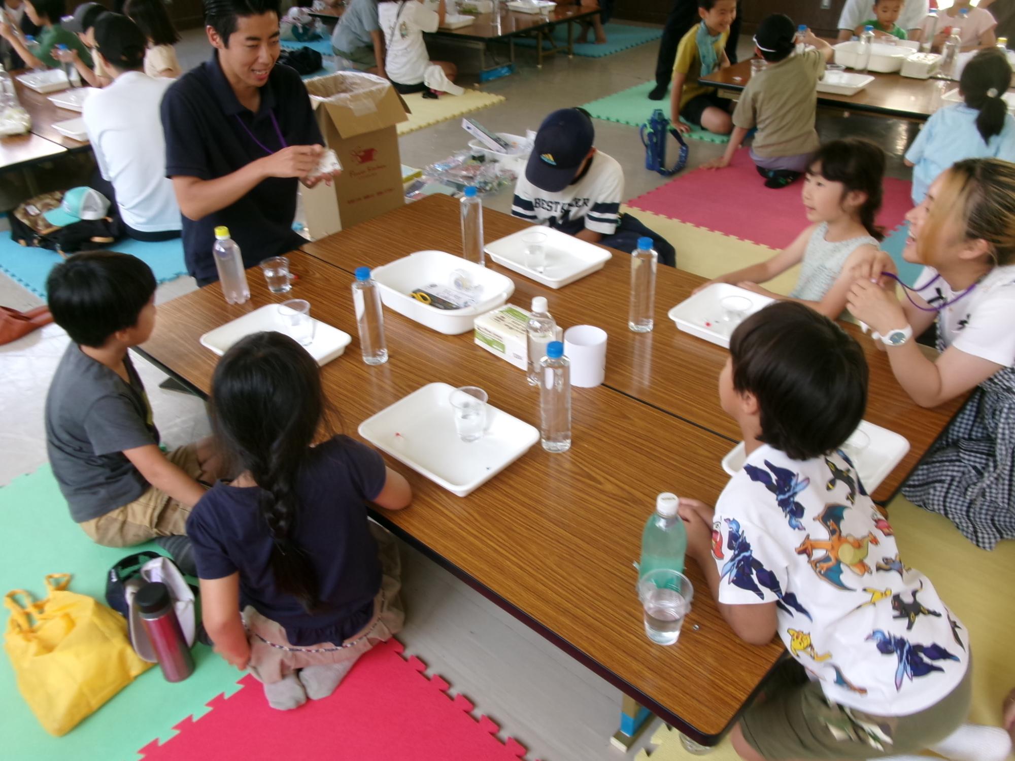 大学生から、浮沈子の作り方の説明を聞いている小学生や保護者の様子の写真