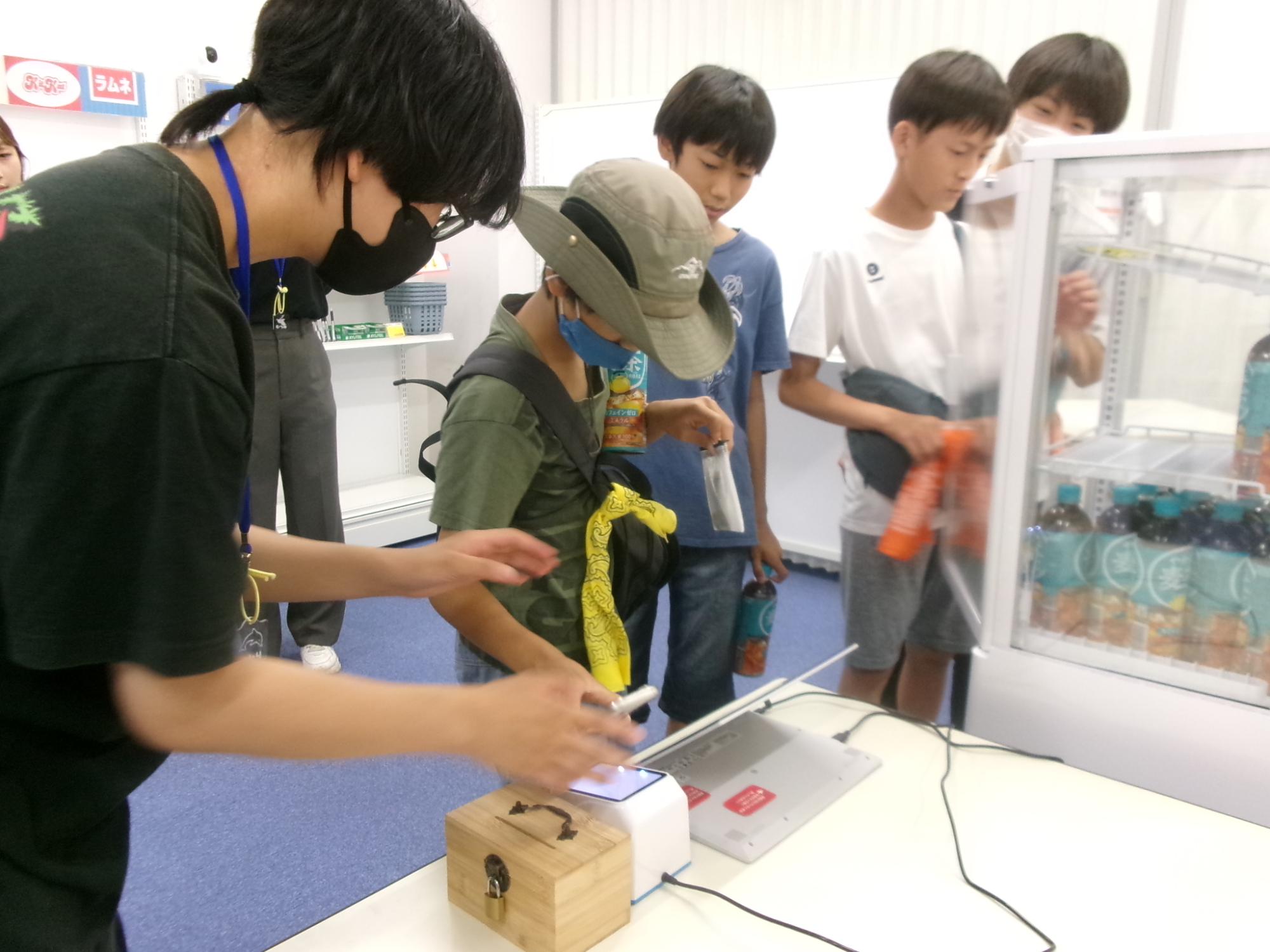 参加児童が、大学生が作製した無人コンビニでスマートフォンを用いてお茶を購入している様子の写真