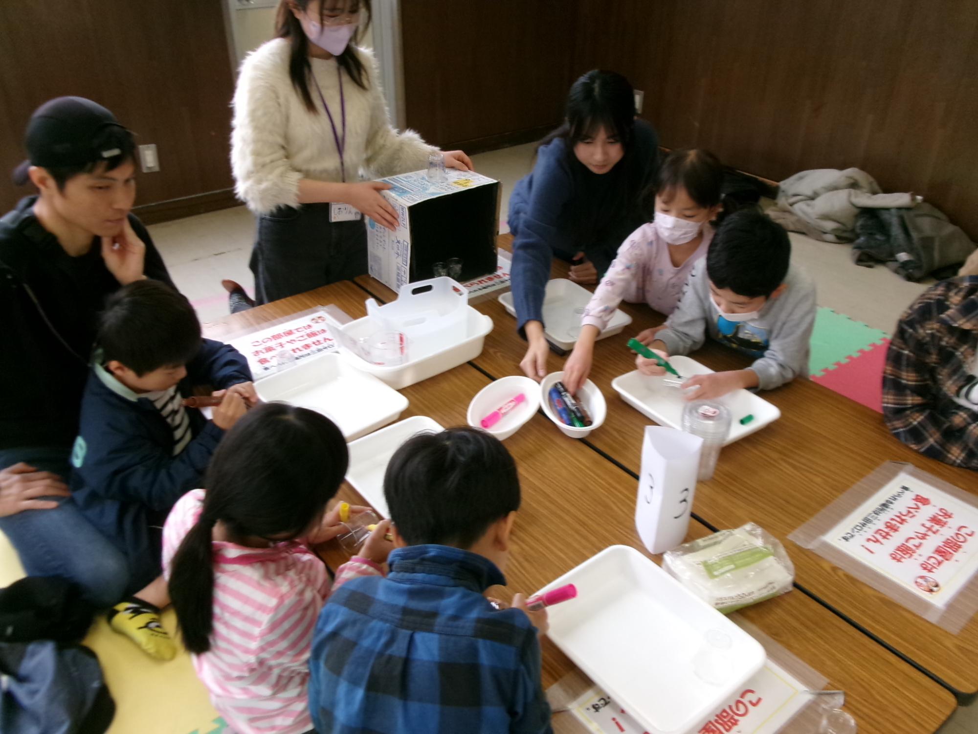 小学生と乳幼児がプラスチックのコップにイラストを描いている様子の写真