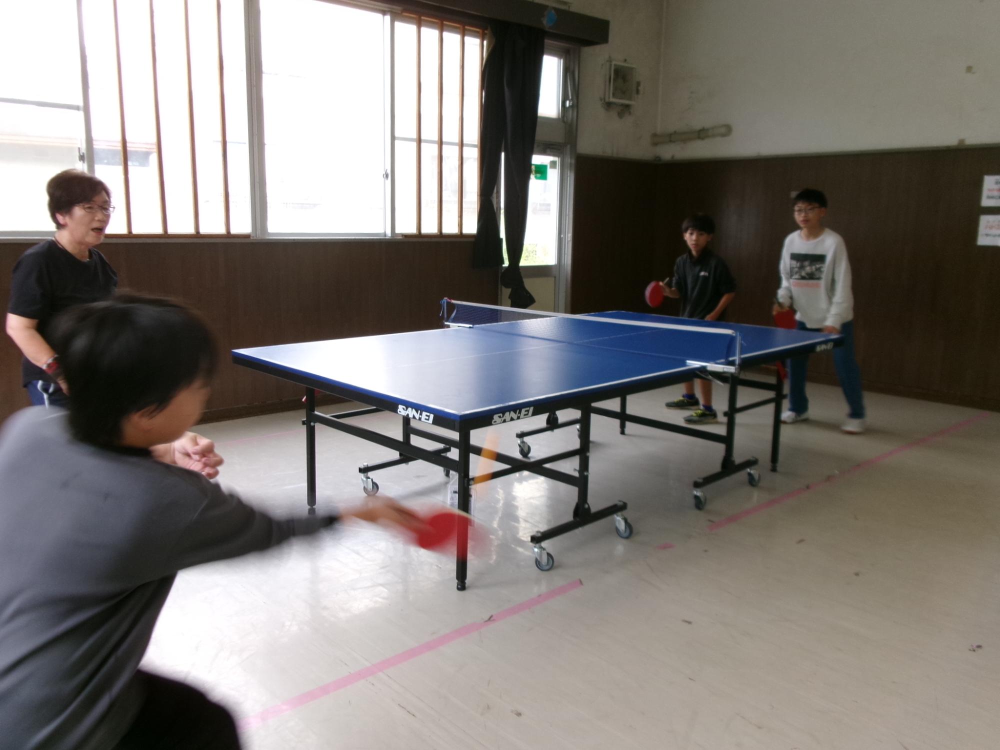 地域住民と中学生と小学生が卓球のダブルスの試合をしている様子の写真