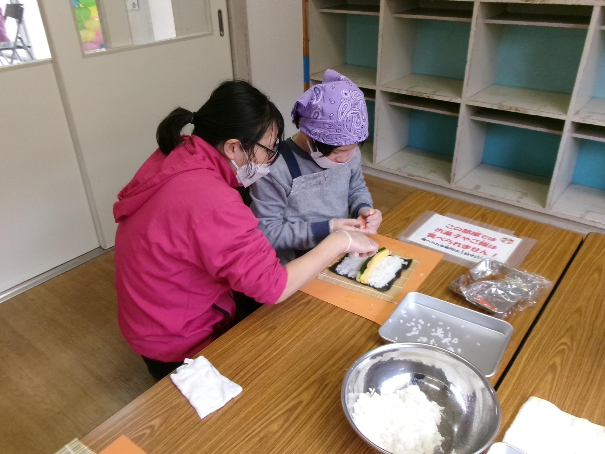 巻きすの上の海苔とお米の上に具材を乗せている小学生親子の写真