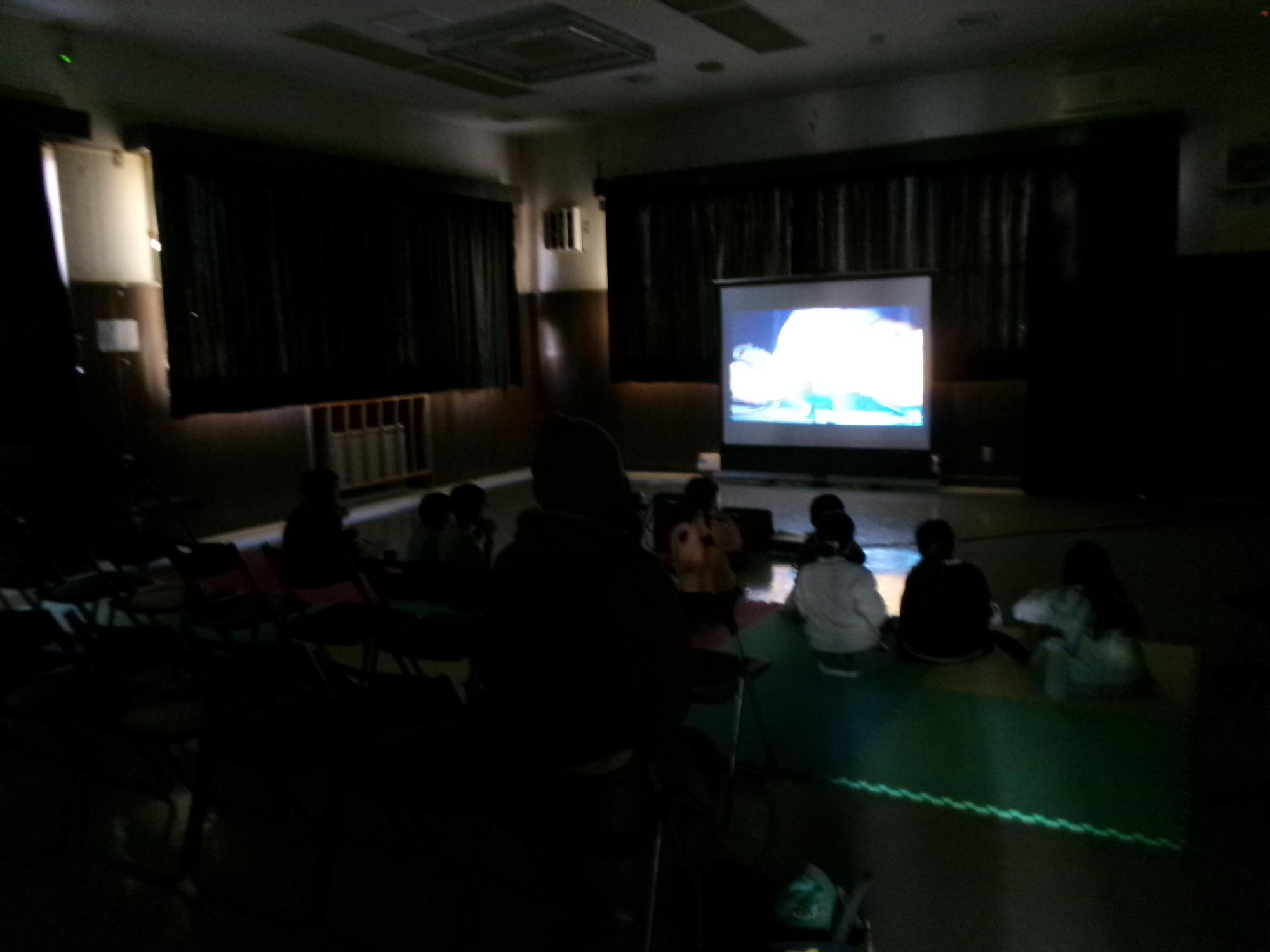 乳幼児親子や小学生が広い部屋で映画を観ながら駄菓子を食べている様子の写真