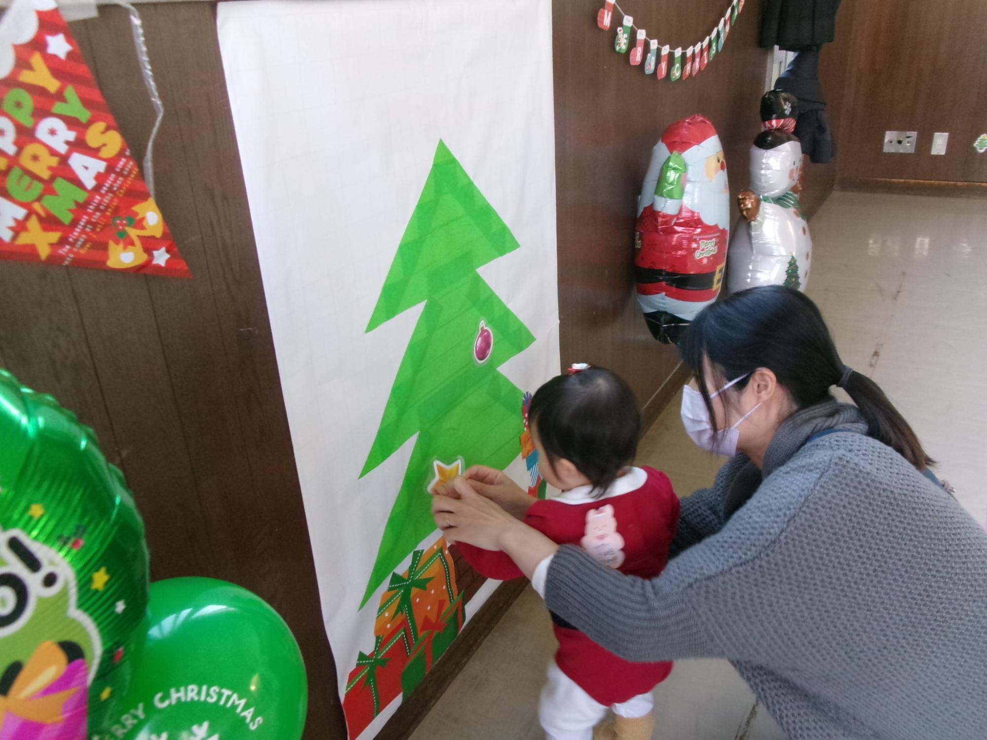 ツリーのポスターに飾りを貼っている幼児の写真