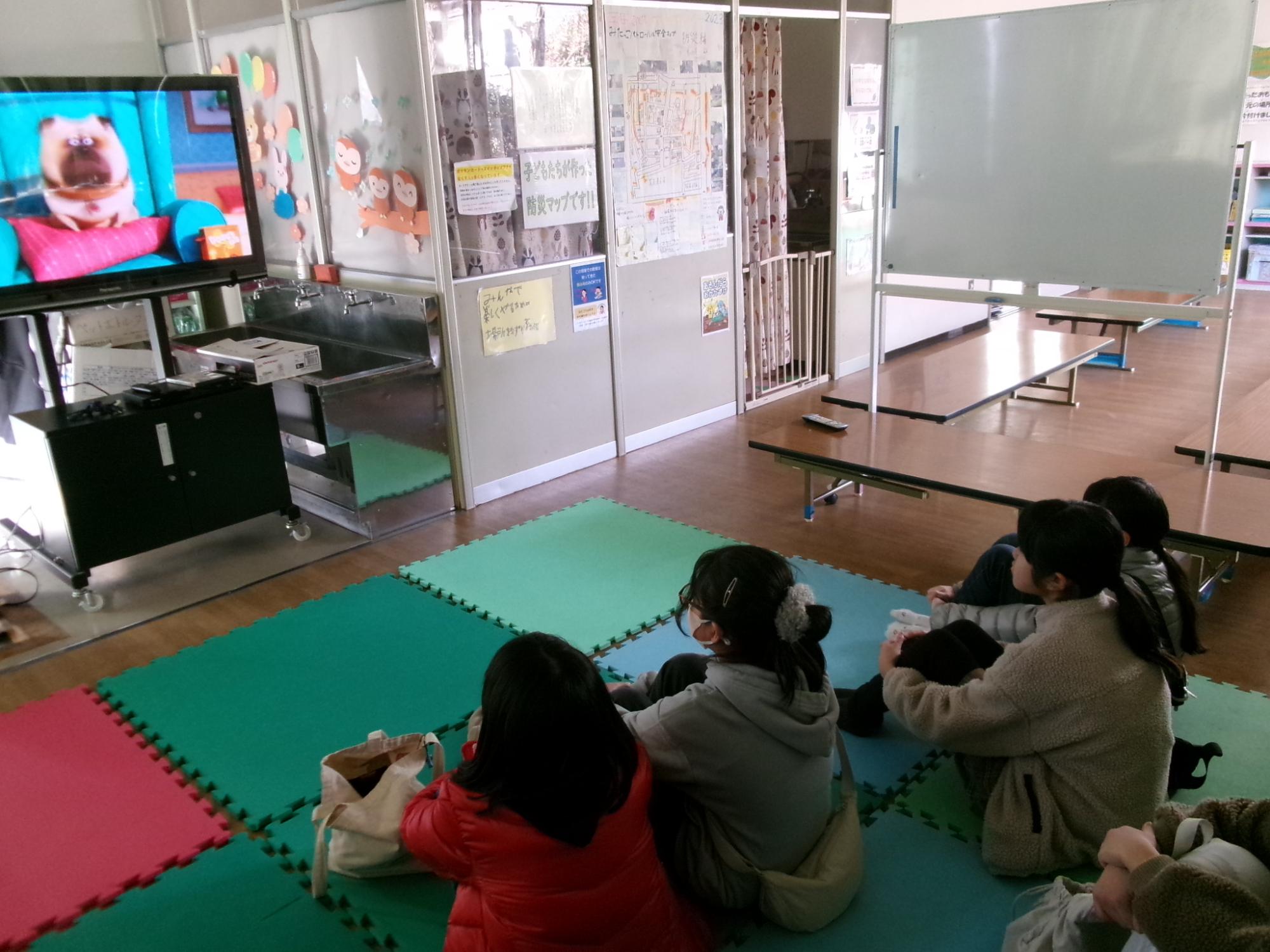 マットの上に座って映画を見る小学生の写真