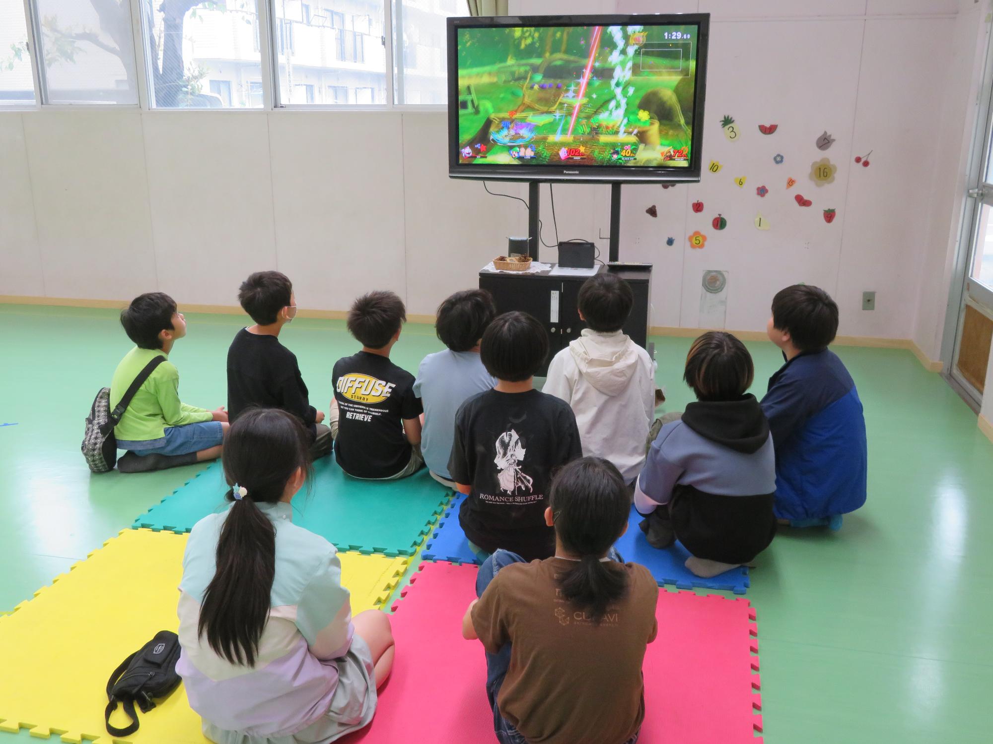 緑色の床の室内で、床に敷かれたマットに座って大きなテレビ画面でゲームをしている子どもたちの写真