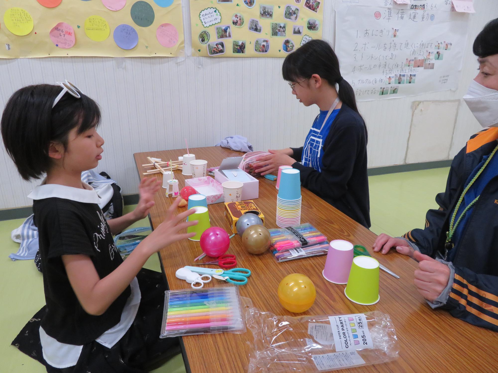 職員と友達に自分の考えた工作を教えている小学生児童の様子