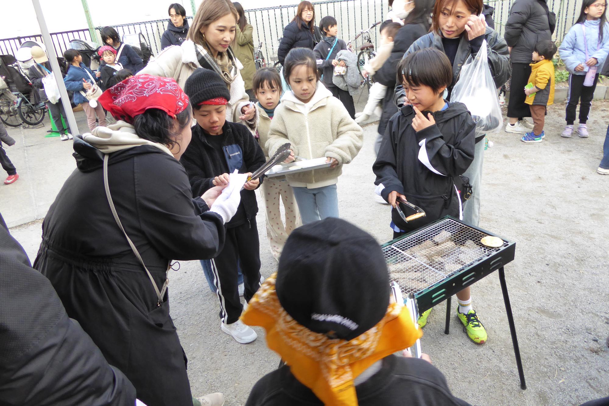 ボランティアの方におせんべいの焼き方を教わる小学生児童の様子の写真