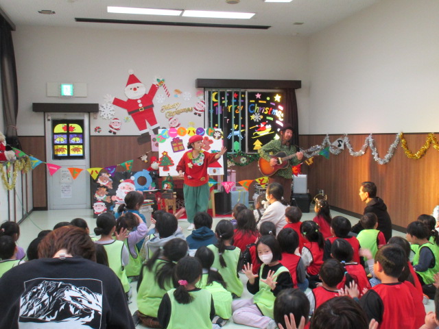 黒色の帽子を被った男性のギター演奏に合わせて、赤い服を着た女性ボーカルが歌を歌っている様子を、子供たちが聞いている後ろ姿の写真