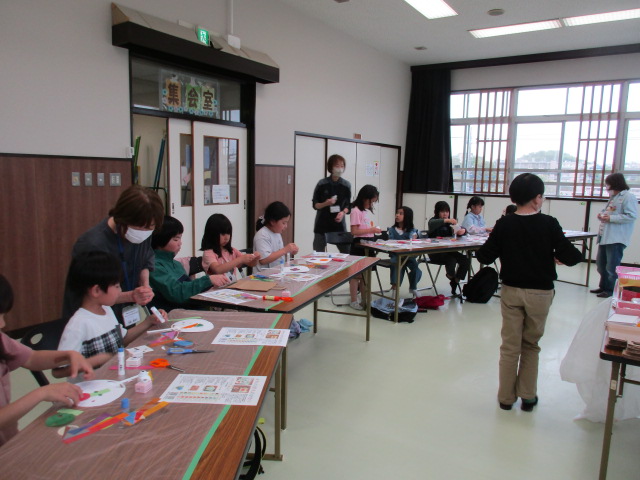 黄緑色の床の室内で、前方に立っている講師の方と、扇状に並べられた机の前で椅子に座っている子どもたちの写真