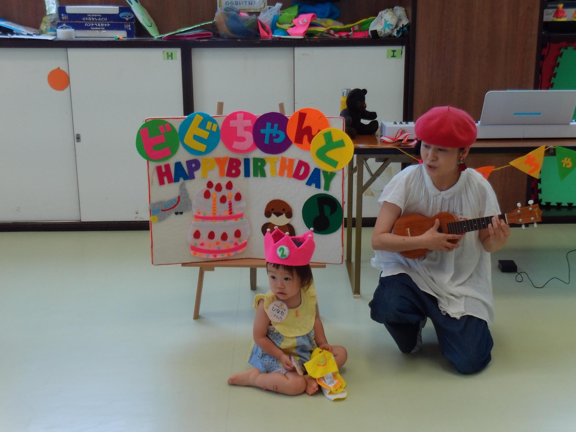 誕生日の装飾がされたボードの隣で演奏をするウクレレ奏者とそれを聞く乳幼児親子の写真