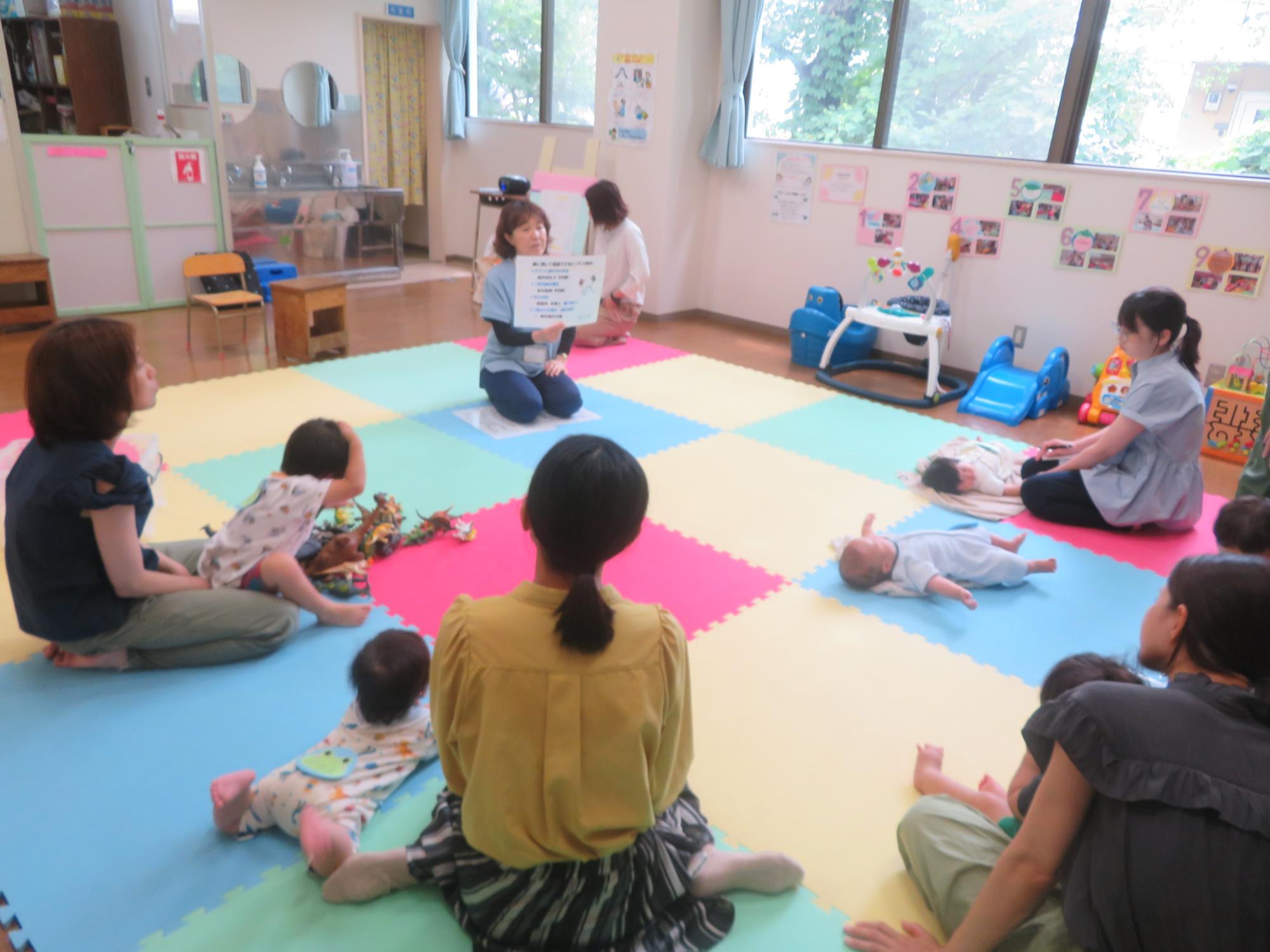 はいはいしながら向かい合う2人の乳幼児の後ろで見守る母親の写真