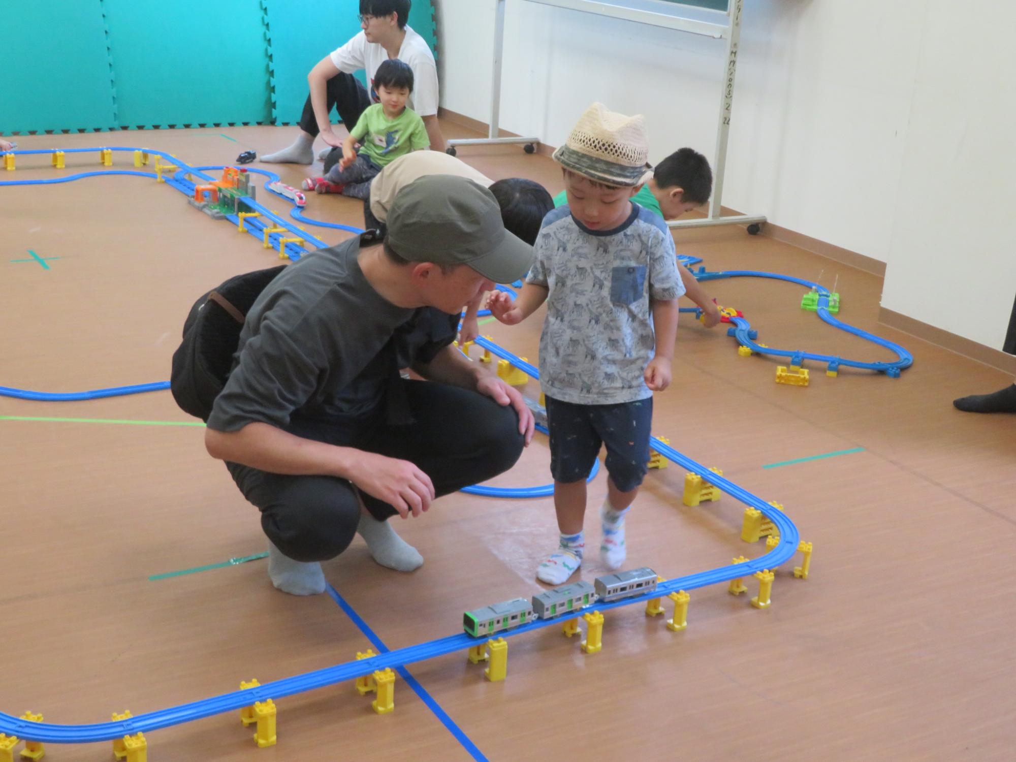 プラレールのコースで電車を走らせて遊んでいる幼児親子の写真