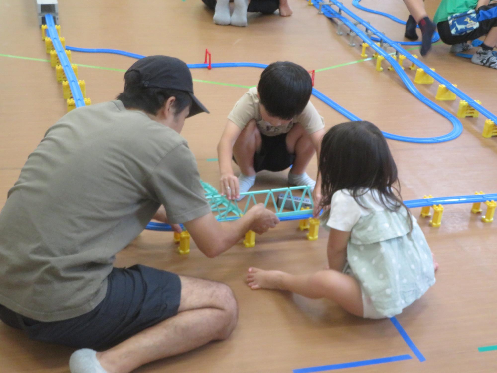 親子3人でプラレールのコースで電車を走らせて遊んでいる写真