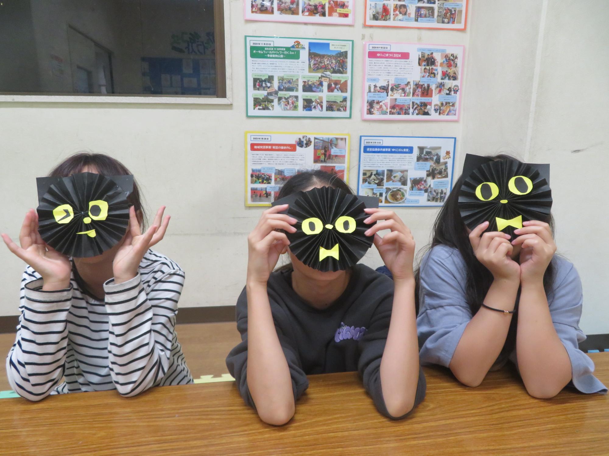ペーパーファンで黒ネコを作ってお面にしている女の子たちの写真
