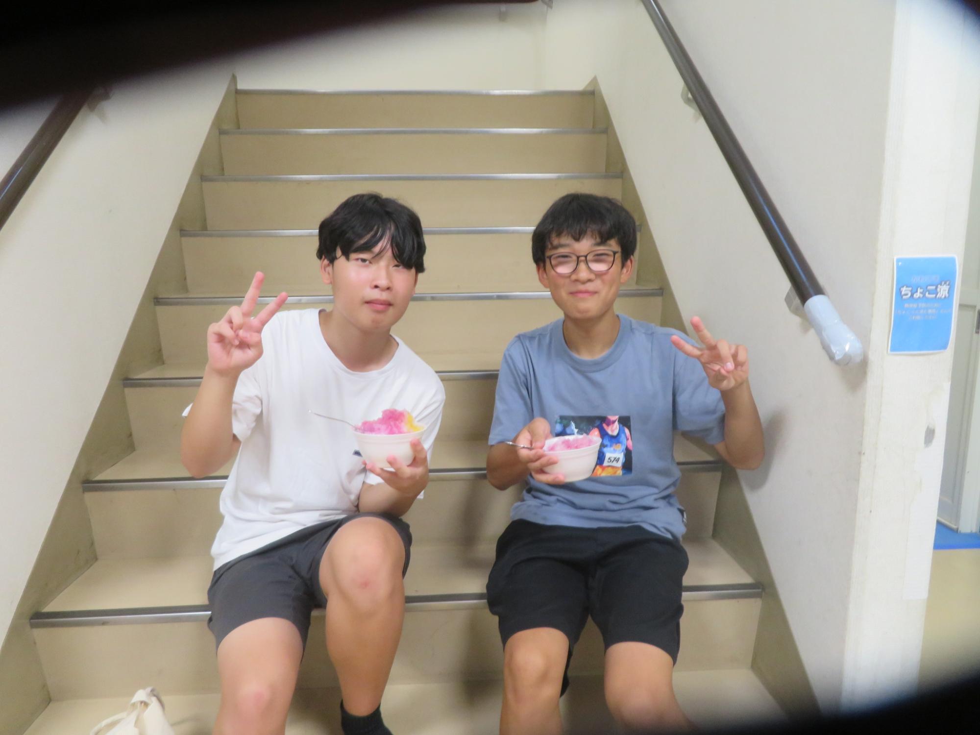 中学生男子2名が階段に座りかき氷をもってピースサインをしている写真